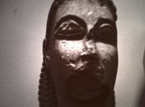 Statue en pierre d'un visage humain avec des traits stylisés et des cheveux tressés.